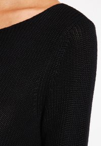 Svart stickad tröja med detaljer som ribbad textur, bred halsringning och åtsittande långa ärmar. Materialet verkar mjukt och elastiskt.