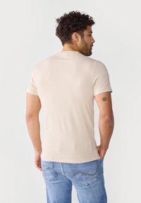 Beige, rövid ujjú póló sima textúrával, testhez álló fazonnal és kerek nyakkivágással, világoskék farmernadrággal párosítva, amelyen hátul zsebek varrása található.