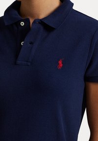 Tmavě modrá polo tričko vyrobené z texturované tkaniny. Má klasický límec, zapínání na knoflíky a červené vyšívané logo na levé straně hrudi.