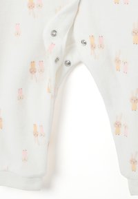Monito blanco hecho de un tejido suave que presenta un patrón repetido de pequeños conejitos estilo caricatura en beige claro y rosa pastel. Con cierres a presión.