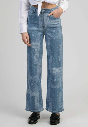 Femme portant un jean taille haute bleu avec un motif patchwork blanc paisley, un chemisier court blanc et des mocassins noirs, debout devant un fond uni.