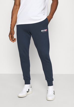 Man draagt marineblauwe joggingbroek met Tommy Jeans-logo op linkerdij, wit T-shirt en witte sneakers met zwarte accenten.