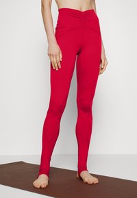 Vysoké červené legíny vyrobené z elastického materiálu, s nařaseným designem v pase a diagonálními švy.