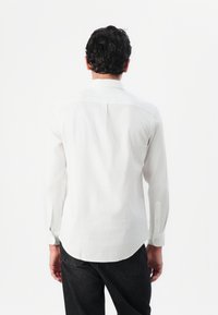 Camisa blanca de manga larga con cuello estándar, pliegue trasero, dobladillo curvado y mangas abotonadas, hecha de una tela ligera.
