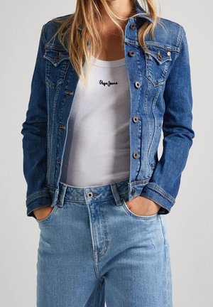 Femme portant une veste en jean bleue par-dessus un débardeur blanc côtelé avec le texte « Pepe Jeans », associé à un jean taille haute bleu clair, mains dans les poches.