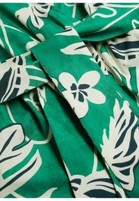 Tissu vert avec un motif de feuilles et de fleurs tropicales blanches et noires, présenté en gros plan avec des plis et des sections qui se superposent.