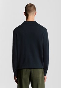 Polo a maniche lunghe di colore blu scuro con colletto, realizzato in tessuto liscio, abbinato a pantaloni verde oliva, con un design minimalista.