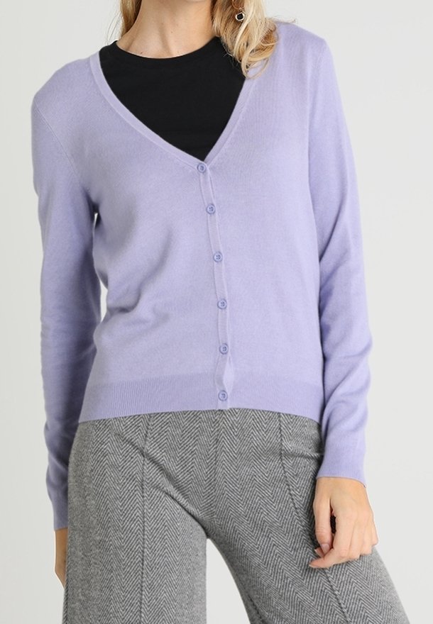 Femme portant un cardigan boutonné violet clair sur un haut noir et un pantalon gris à motif chevrons, debout avec une main posée sur la cuisse.