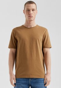 Brązowy t-shirt bawełniany z okrągłym dekoltem i krótkimi rękawami, o gładkiej fakturze. Połączony z dżinsami, wyeksponowany na szarym tle.