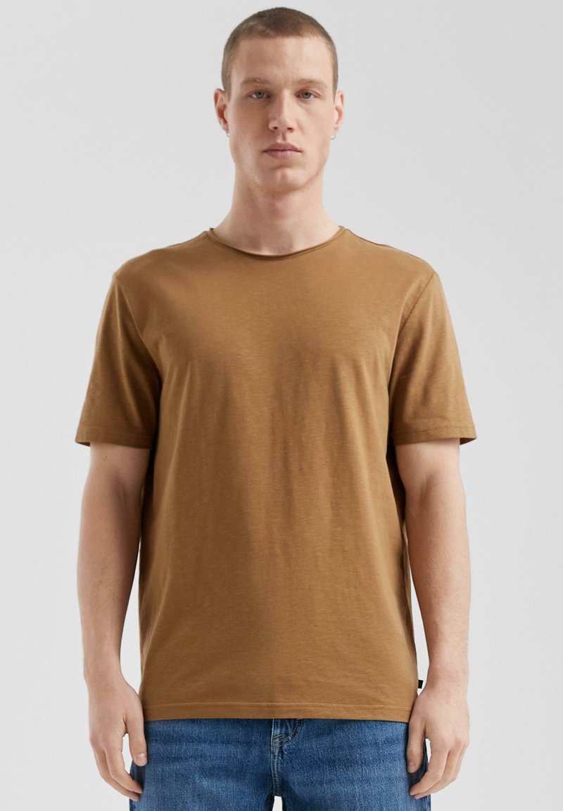 Brązowy t-shirt bawełniany z okrągłym dekoltem i krótkimi rękawami, o gładkiej fakturze. Połączony z dżinsami, wyeksponowany na szarym tle.