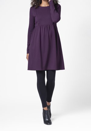 Donna con un vestito viola a maniche lunghe, leggings neri e stivaletti neri, in piedi davanti a uno sfondo bianco.