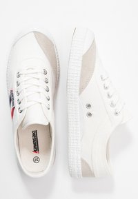 White canvas sneakers featuring beige suede accents and textured rubber soles. Six eyelets and a small brand tag with red and blue details.