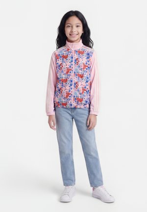 Mladá dívka stojící, oblečená v růžové květinové zipové bundě, světle modrých džínách a bílých teniskách, na jednobarevném bílém pozadí.