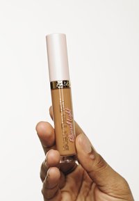 Correcteur liquide dans un tube cylindrique transparent avec un capuchon rose. L'étiquette indique "BORN THIS WAY Ethereal Light" en texte doré et rose.