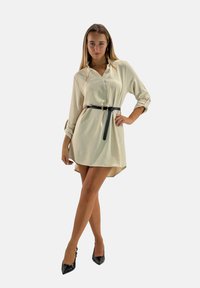 Vestito camicia beige con bottoni, maniche lunghe, con cintura in vita in pelle nera. Presenta un orlo asimmetrico e una texture liscia.