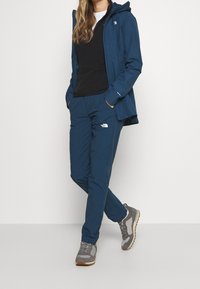 Marinfärgad vattentät jacka med huva, svart skjorta och matchande marinfärgade byxor. Grå och vita sportskor. Har The North Face-logotyper på byxorna.