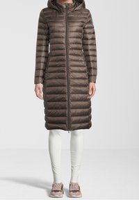 Ruskea puffatakki hupulla, jossa on pystysuora tikkikuvio, täyspitkä vetoketju ja sivutaskut. Puettu valkoisten legginsien ja lenkkareiden päälle.