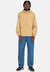 Beige letvægts hoodie-jakke med elastiske manchetter, sidelommer og logo-detalje. Kombineret med afslappede lyseblå jeans og sneakers.
