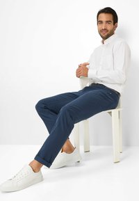 Weißes Button-Down-Hemd, schmal geschnittene navyblaue Hose und weiße Sneaker. Die Hose hat aufgerollte Säume, das Hemd verfügt über eine Brusttasche.