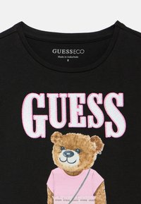 Μαύρο βαμβακερό T-shirt με καρτούν αρκούδα καφέ γούνας, ροζ μπλούζα και ασημένια αλυσίδα. Το "GUESS" είναι κεντημένο σε ροζ και λευκό χρώμα.