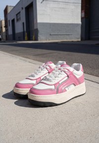 Paire de baskets blanches et roses à lacets avec des semelles épaisses placée sur un trottoir en béton près d'une rue et des bâtiments.