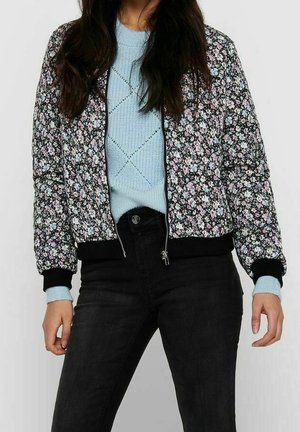 Person trägt eine schwarze Jacke mit floralem Muster und Reißverschluss über einem hellblauen Strickpullover mit schwarzen Hosen vor einem einfarbigen Hintergrund.