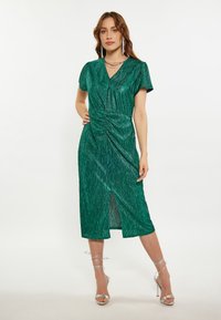 Rochie verde, de lungime medie, cu dungi verticale subtile, mâneci scurte tip puff, stil înfășurat, talie fronsată și deschidere frontală. Asortată cu tocuri argintii.