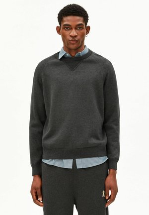 Homme debout devant un fond uni, portant un pull gris foncé sur une chemise à col rayée bleue et un pantalon gris foncé assorti.