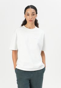 White cotton t-shirt with a relaxed fit, short sleeves, and a blue logo printed on the left chest. Paired with dark green denim trousers.