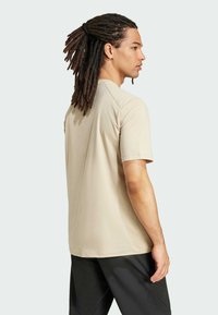 Beige short-sleeve t-shirt made of smooth, lightweight fabric. Features a relaxed fit with simple design, worn with black pants, seen from the back.