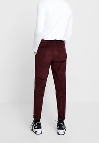 Mörkröda slim-fit byxor med texturerat tyg, med bakfickor och en avsmalnande benform, kombinerade med svarta och vita sneakers.