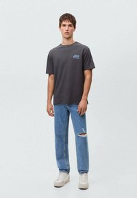 Gray short-sleeve t-shirt with “Fuel Garage” graphic, paired with blue distressed jeans and beige sneakers, standing against a light background.