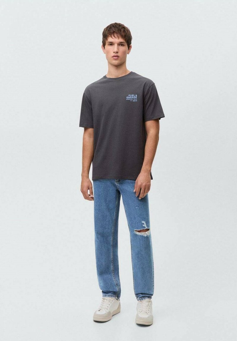 Gray short-sleeve t-shirt with “Fuel Garage” graphic, paired with blue distressed jeans and beige sneakers, standing against a light background.