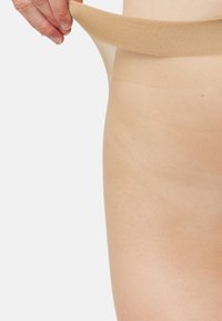 Beige gennemsigtige strømper med et blødt, bredt elastikbånd, der bliver trukket op, hvilket fremhæver en glat tekstur mod lys hud.