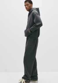 Ung man som bär en mörkgrå hoodie och matchande sweatpants, står på sidan med händerna i fickorna och har svarta sneakers med krämfärgade snörningar.