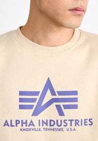 Beige szövetszitter ribbelt nyakkal, amelyen egy lila grafikai design látható egy 'A' betűvel és vízszintes csíkokkal, az "ALPHA INDUSTRIES" felirattal.