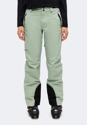 Modèle portant un pantalon de ski vert pâle avec des empiècements noirs dans le bas des jambes, un haut noir et des chaussures de ski noires sur un fond uni.