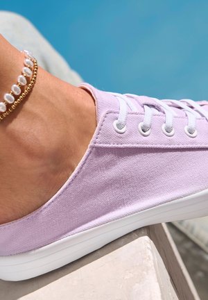 Light purple canvas shoe with white laces and sole, featuring a textured surface. An ankle bracelet with pearls is visible.