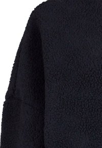 Sweatshirt en polaire noire avec une surface douce et texturée. Présente une silhouette carrée et des épaules tombantes pour un ajustement décontracté.