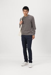 Grauer gestrickter Pullover mit Rundhalsausschnitt, marineblaue Slim-Fit-Hosen und weiße Sneakers. Das Modell hält eine beige Tragetasche. Einfaches Design, minimale Details.