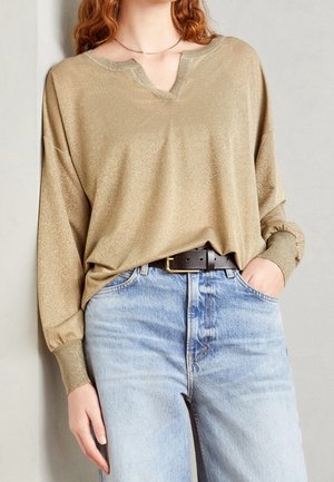 Persona con un top dorado suelto y brillante de manga larga con escote en v, jeans de tiro alto en azul claro y cinturón negro.