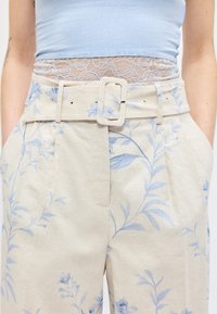 Γυναίκα φορώντας μπεζ ψηλόμεσο φλοράλ παντελόνι με ζώνη και ανοιχτό μπλε τοπ, που είναι μερικώς τακτοποιημένο μέσα στη δαντελένια μέση.