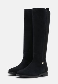 Par de botas de gamuza negras hasta la rodilla con paneles laterales elásticos y tacones bajos, que cuentan con un pequeño logo metálico en el lado exterior cerca del tobillo.