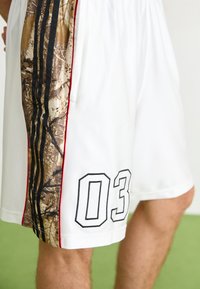 Shorts de sport blancs avec un panneau latéral en imprimé camouflage, comportant trois bandes noires et le numéro "03" en détail brodé noir.