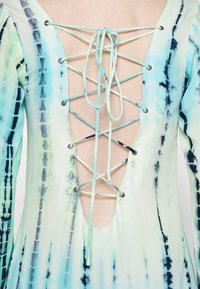 Šaty s vázáním na zádech, které mají hluboký výstřih do V, vyrobené z lehkého materiálu s modro-zeleným tie-dye vzorem a stříbrnými očky.