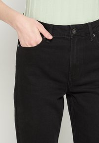 Melnu denimu bikses ar šauru piegriezumu, ar priekšējo kabatu, pogu aizdari un smalkiem šuvju detaļām. Materiāls ir gluds uz tausti.