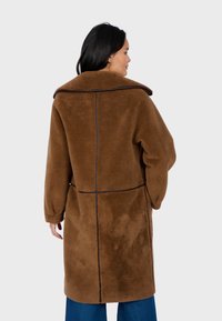 Cappotto in pile marrone con colletto alto, spalle scese e dettagli delle cuciture a contrasto. La parte posteriore è liscia e ha una vestibilità rilassata.