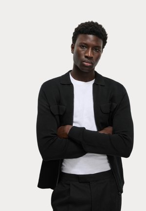 Black zip-up cardigan with a collar, featuring two chest pockets. Worn over a white t-shirt, textured knit with ribbed cuffs.