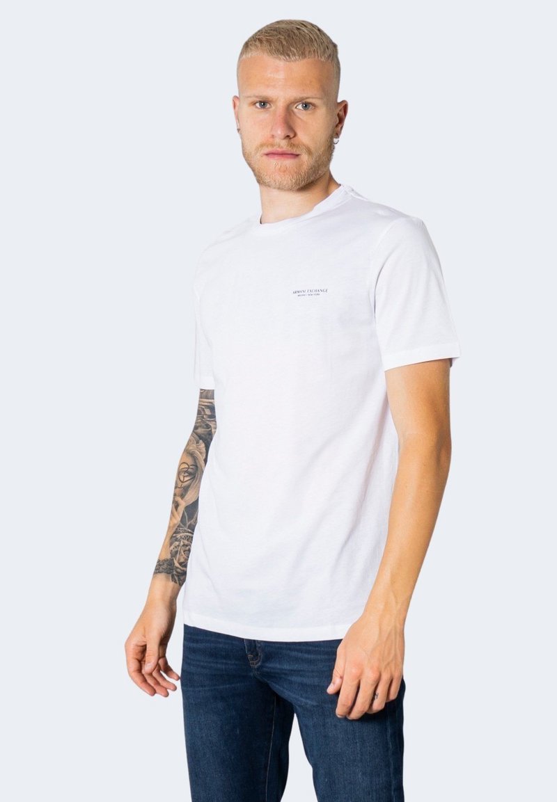 Tricou alb din bumbac cu mâneci scurte, guler rotund și un logo subtil pe piept. Modelul poartă blugi din denim albastru.