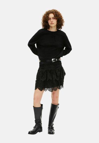 Mujer con cabello rizado, vestida con un suéter negro, falda de encaje negro con cinturón y botas de cuero negro hasta la rodilla, de pie contra un fondo blanco.
